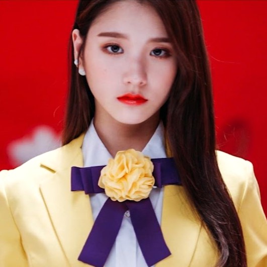 Heejin from the 'Vivid' music video. She is staring at the viewer. She has long brown hair and is wearing a yellow outfit.